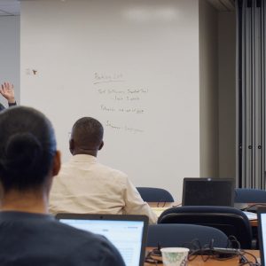 Photo: Trainer standing in front of room talking to rows of trainees sitting at computers.