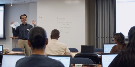 Photo: Trainer standing in front of room talking to rows of trainees sitting at computers.