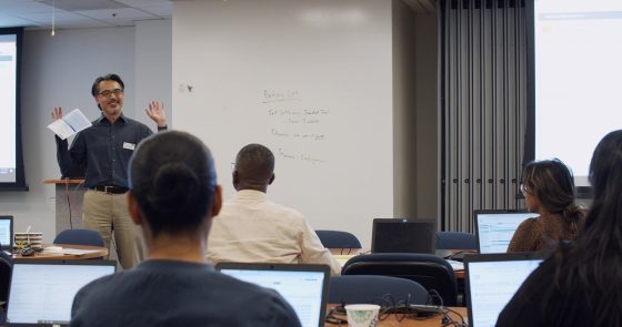 Photo: Trainer standing in front of room talking to rows of trainees sitting at computers.